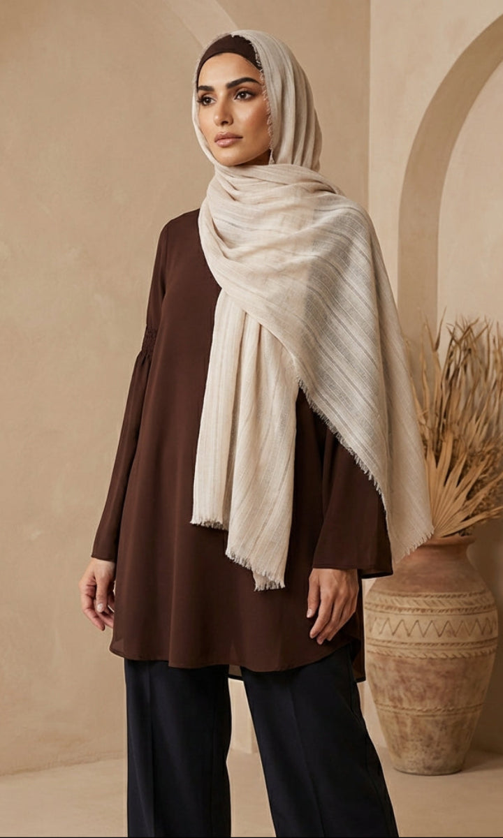 Woman wearing a brown top and black pants with a beige hijab, standing against a beige wall.