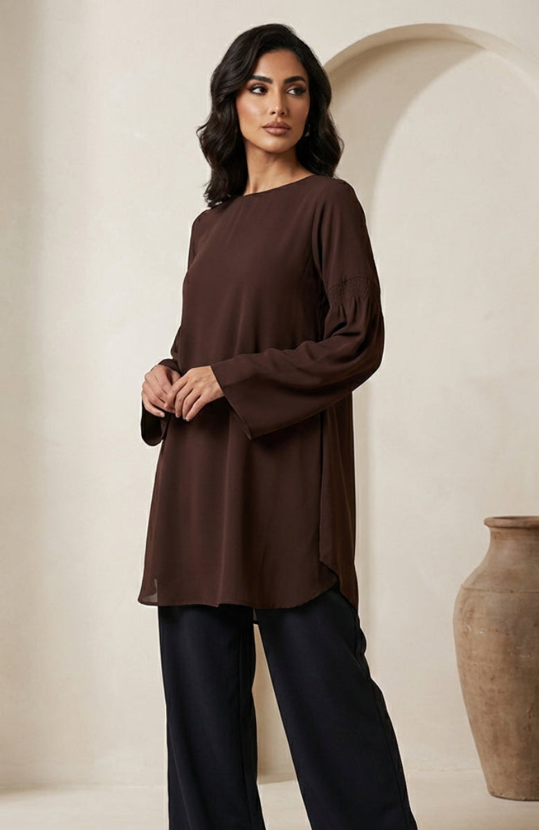 Woman wearing a brown long-sleeve top and black pants standing next to a large vase in a neutral setting.