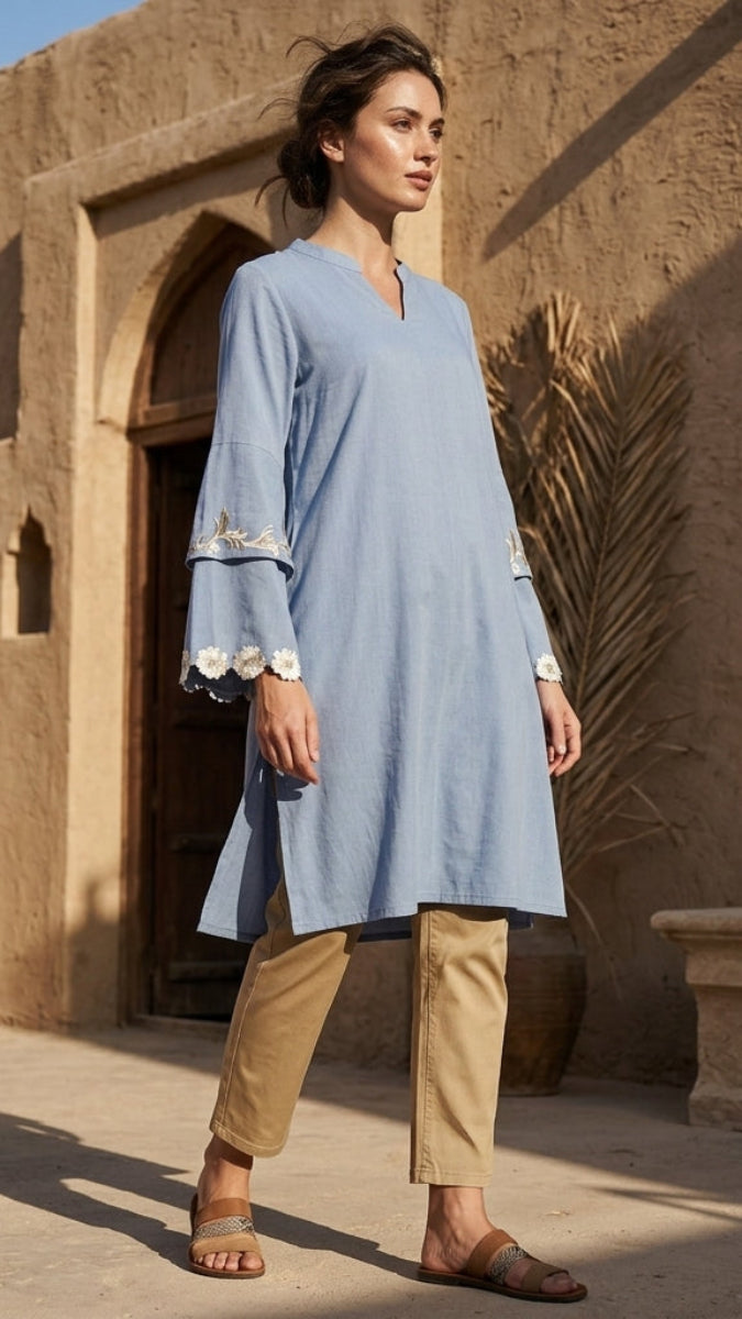 Woman in a light blue dress and beige pants standing in front of a sandy architectural background
