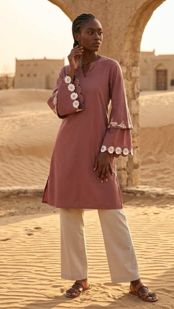 Woman in a pink dress with white floral details standing in a desert landscape.