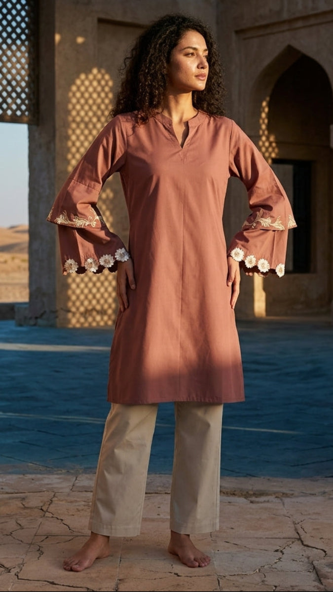 Woman in a long, light brown dress standing in a desert setting with architectural elements.