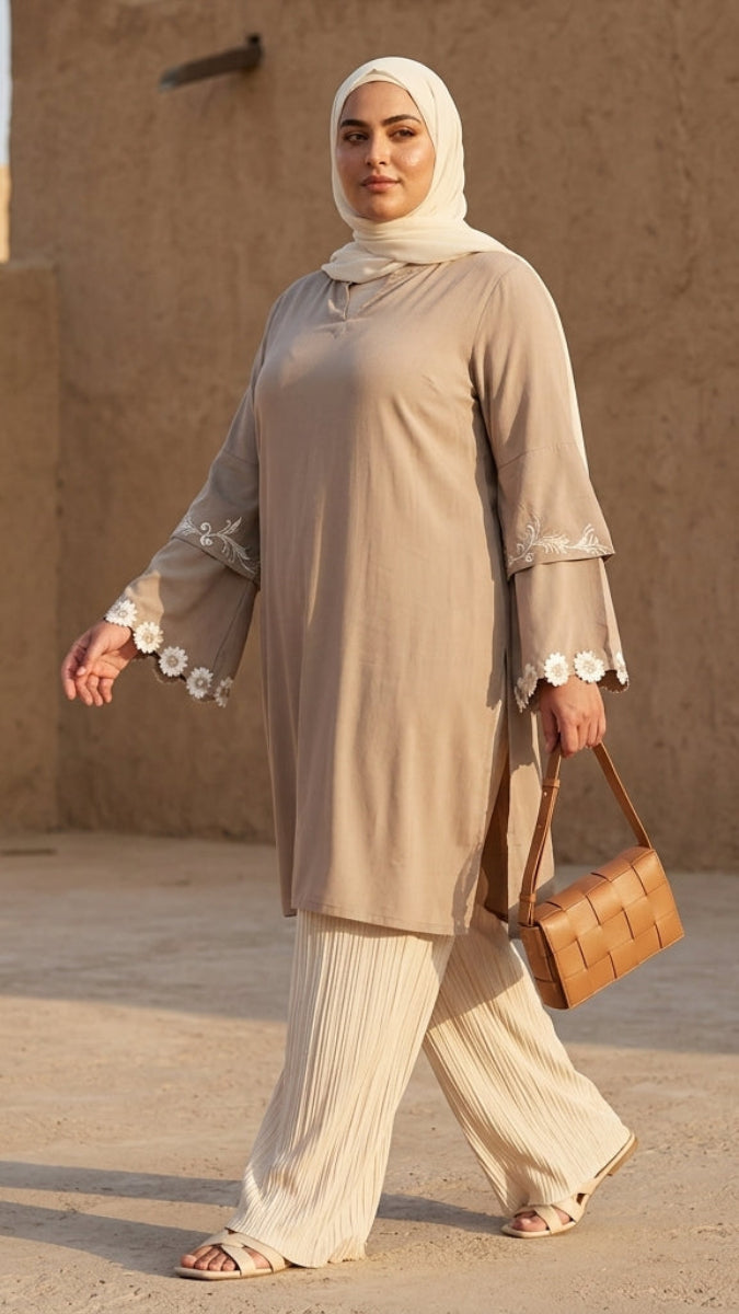 Woman in beige traditional outfit with a headscarf, standing in front of a sandy wall.
