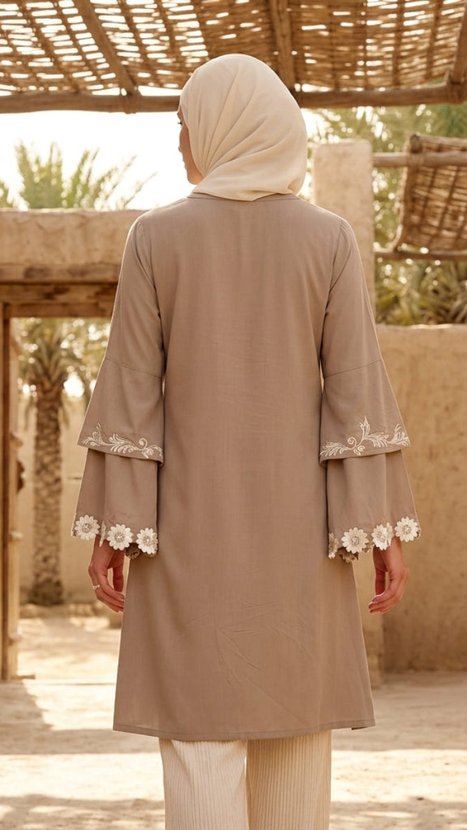 Person wearing a beige dress with floral embroidery in an outdoor setting with stone walls and palm trees.