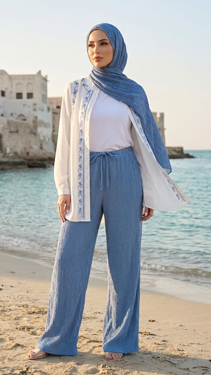 Woman in blue hijab and pants standing on a beach with ocean and buildings in the background