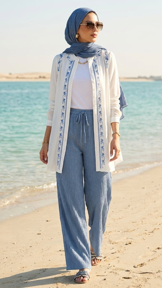 Woman in a hijab and white outfit standing on a beach with clear blue water.