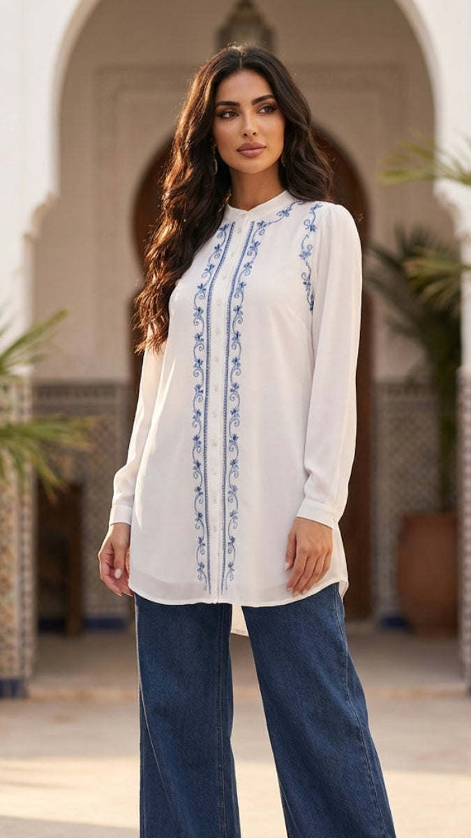 Woman wearing a white blouse with blue embroidery and blue jeans standing in a decorative outdoor setting.