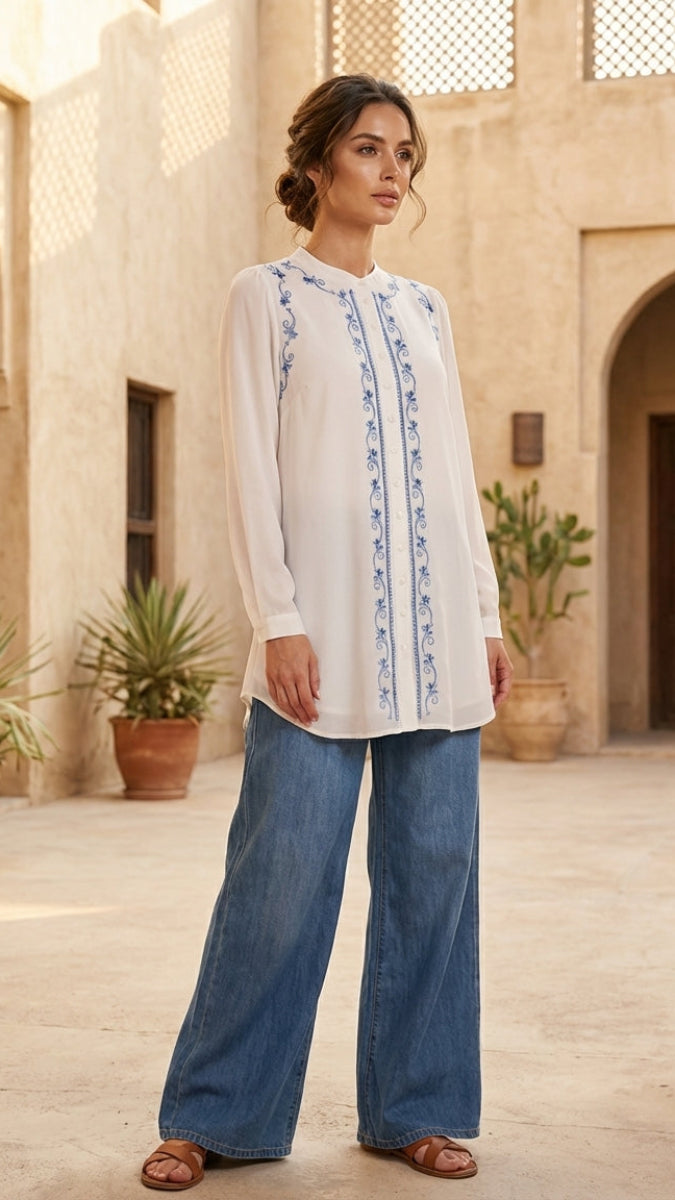 Woman wearing a white blouse with blue embroidery and blue jeans in a courtyard setting.