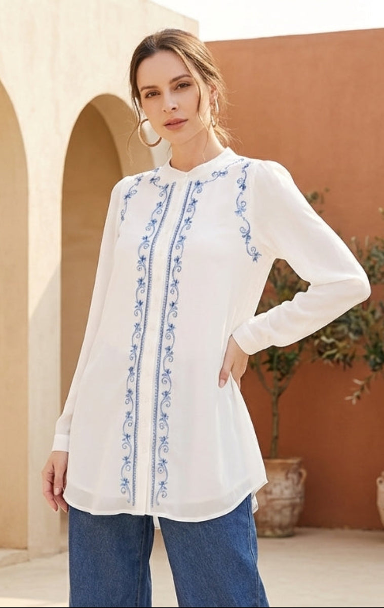 Woman in a white blouse with blue patterns standing in a sunlit courtyard with arches and plants.