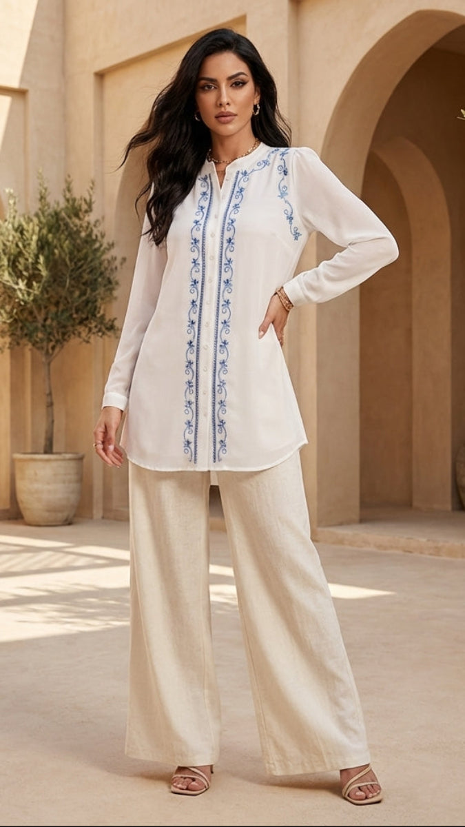 Woman in a white outfit with blue embroidery standing in a sunlit courtyard with arches and potted plants.