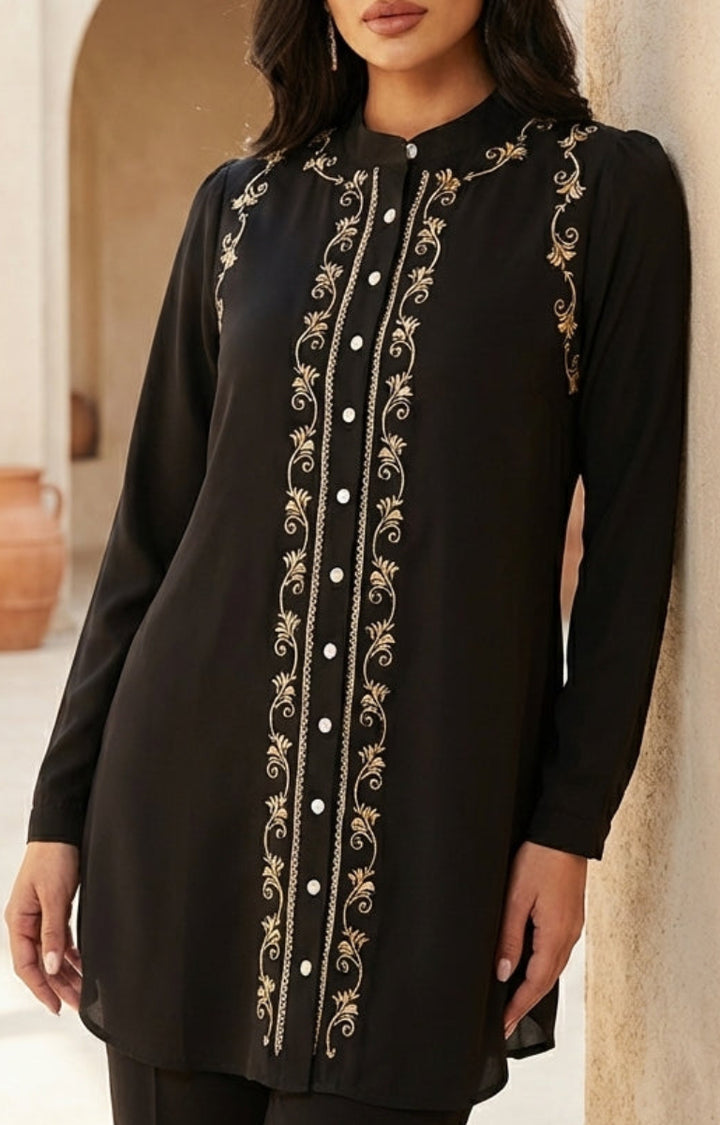 Woman wearing a black embroidered dress in an outdoor setting with beige walls and terracotta pots.