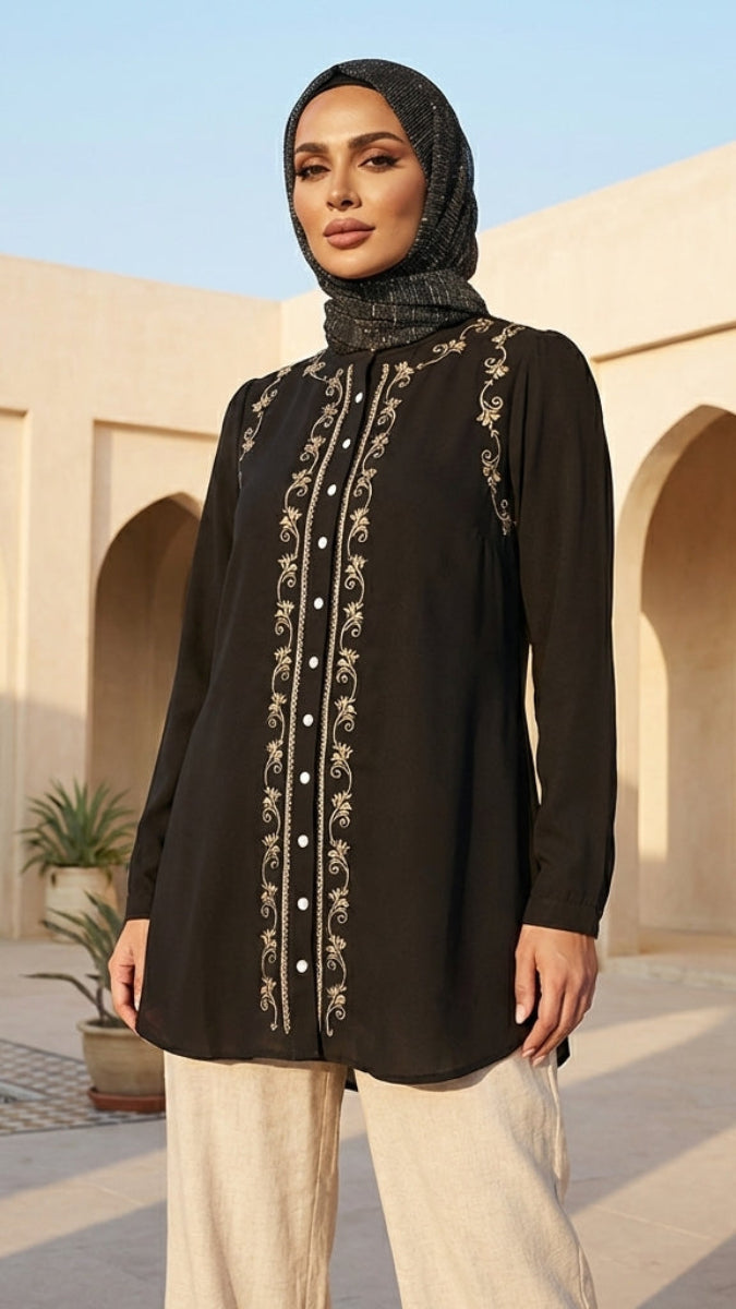 Woman wearing a black embroidered top and hijab in a sunlit courtyard.