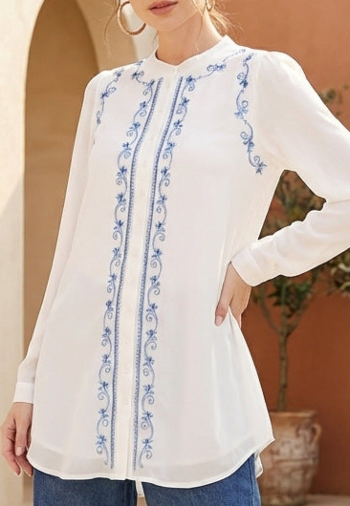 Woman in a white blouse with blue patterns standing in a sunlit courtyard with arches and plants.