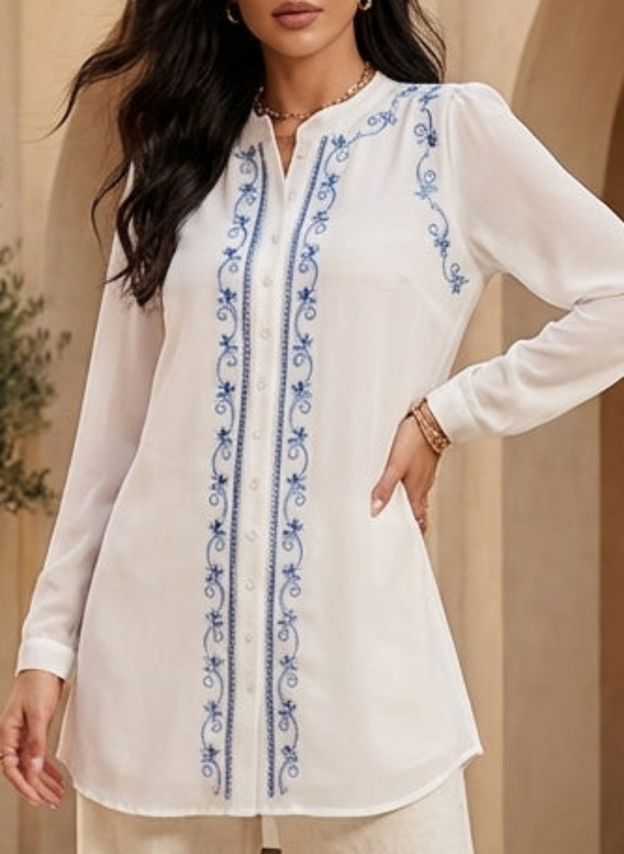 Woman in a white outfit with blue embroidery standing in a sunlit courtyard with arches and potted plants.