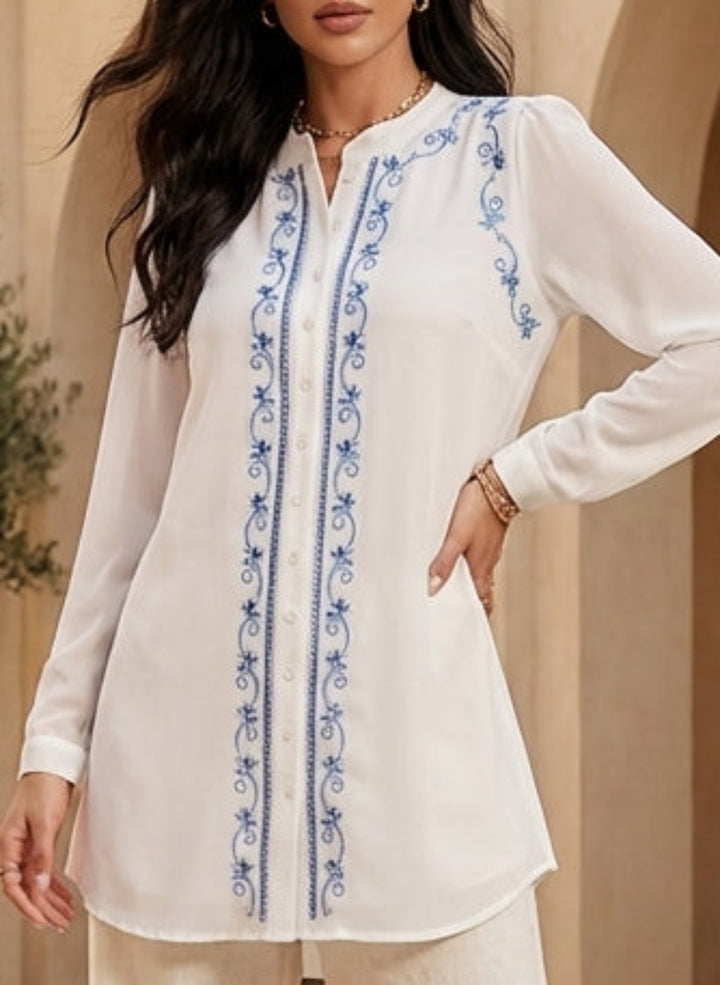 Woman in a white outfit with blue embroidery standing in a sunlit courtyard with arches and potted plants.