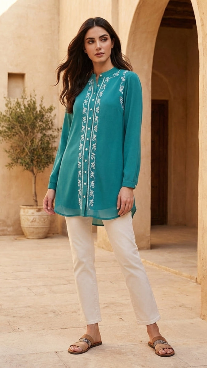 Woman wearing a teal embroidered top and white pants standing in a sunlit outdoor setting with arches and plants.
