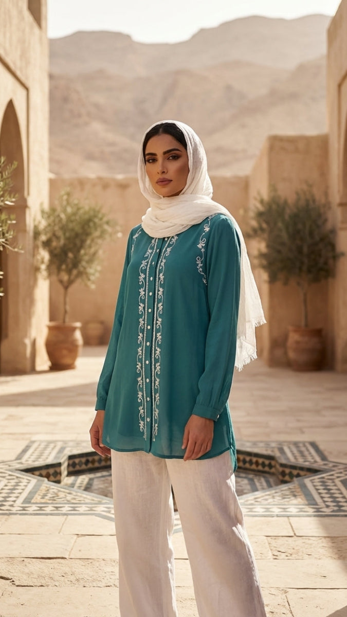 Woman in a teal embroidered top and white hijab standing in a desert courtyard with mountains in the background.