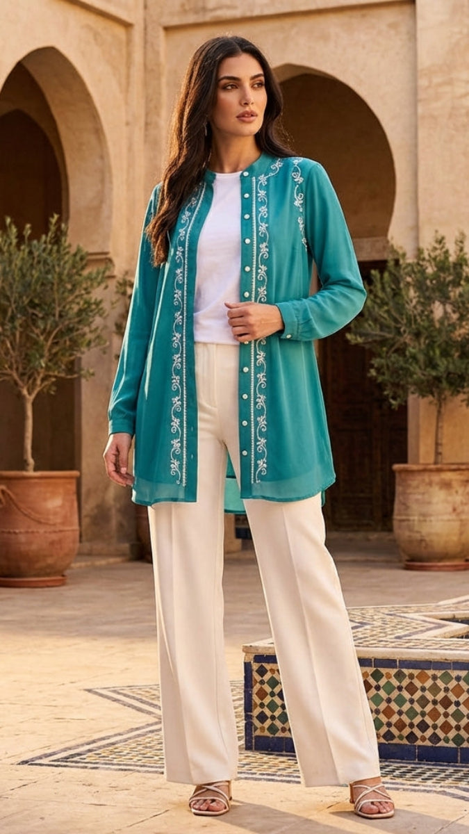 Woman in teal embroidered jacket and white pants standing in a courtyard with arches and potted plants.