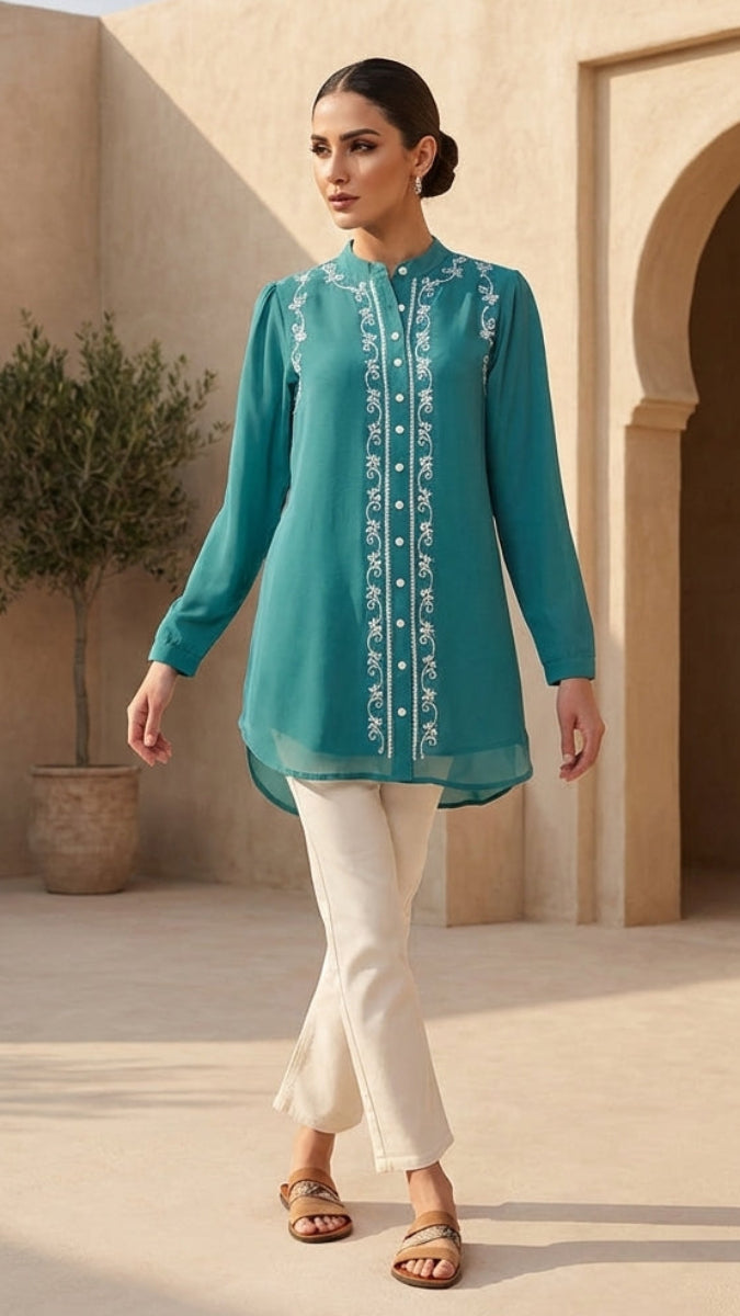 Woman in a teal embroidered top and white pants standing in front of beige architectural elements.