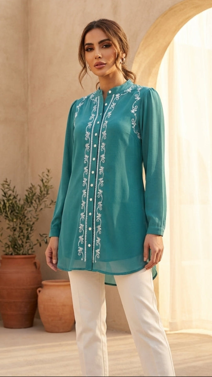 Woman wearing a teal embroidered top and white pants in a warm-toned outdoor setting.