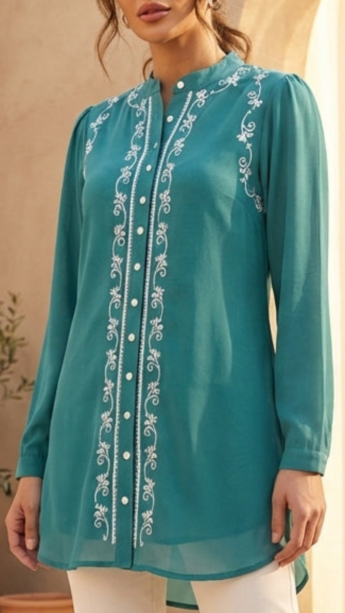 Woman wearing a teal embroidered top and white pants in a warm-toned outdoor setting.