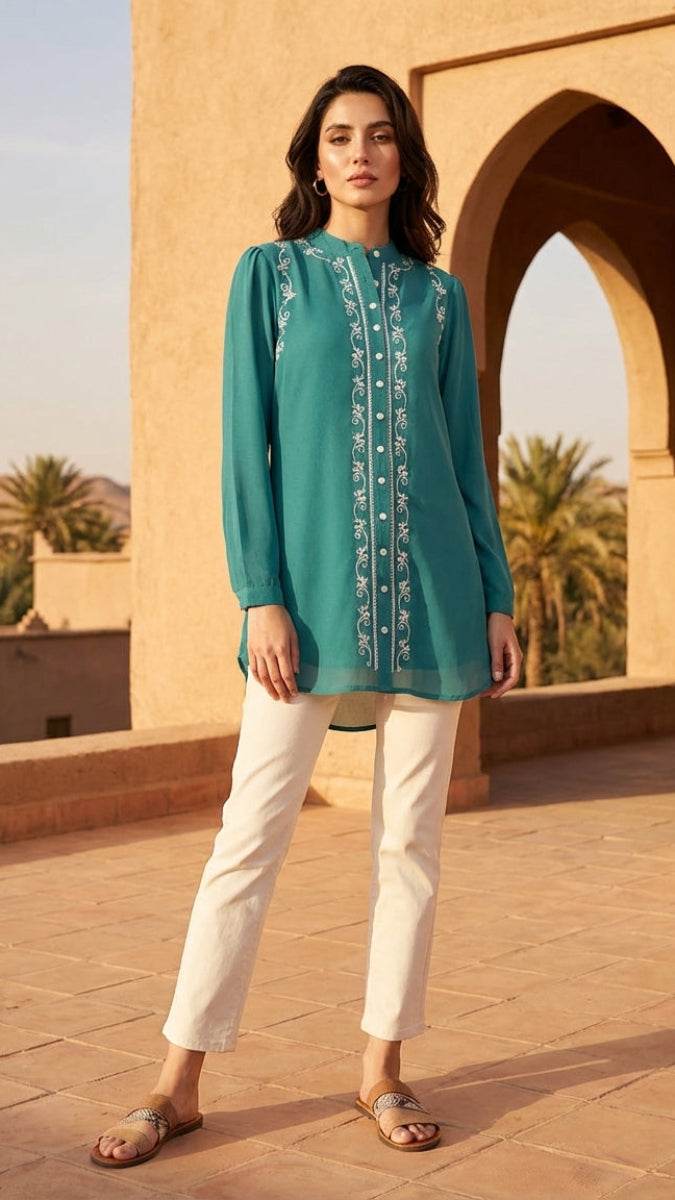 Woman wearing a teal embroidered top and white pants standing in front of an archway with palm trees in the background.