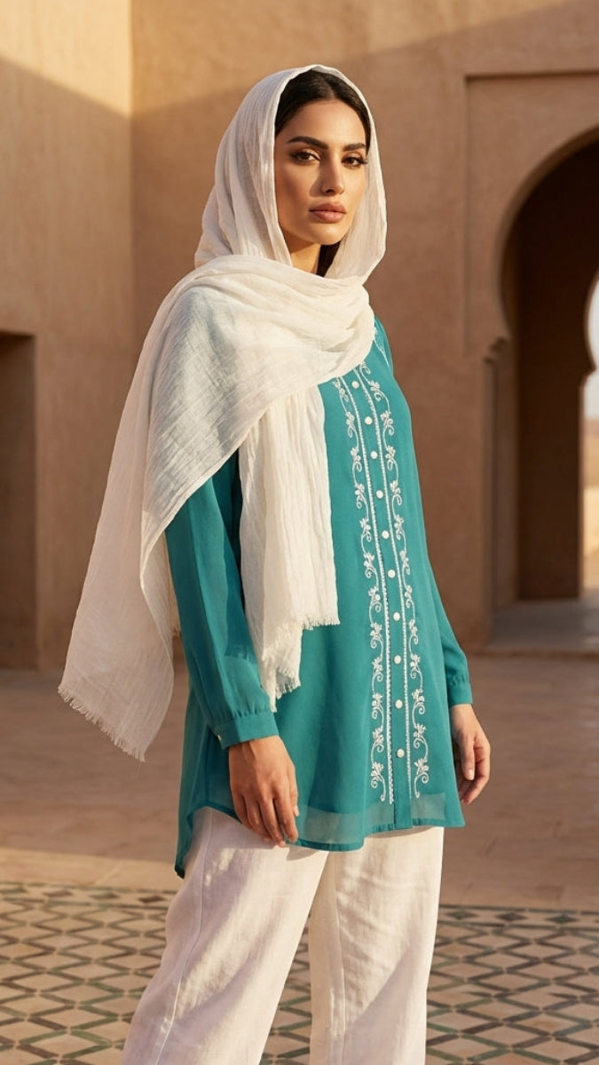 Woman wearing a teal top with white embroidery and a white scarf in a desert setting.