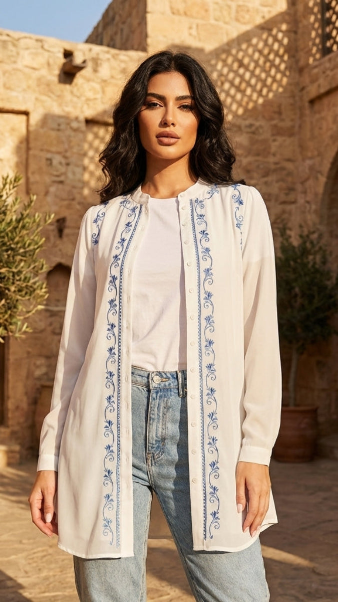 Woman wearing a long, embroidered white cardigan in a sunlit stone courtyard.