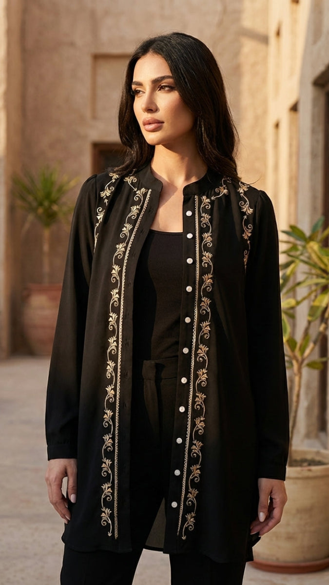 Woman wearing a black embroidered jacket in an outdoor setting with plants and stone walls.