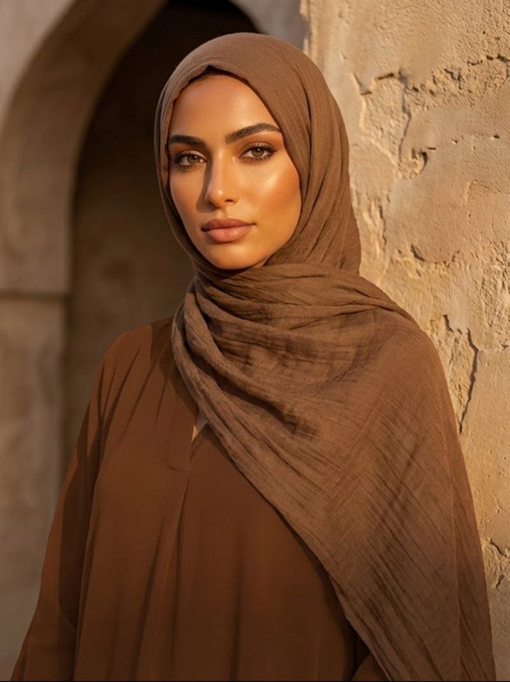 Woman in a brown hijab standing in front of a stone wall with an archway.