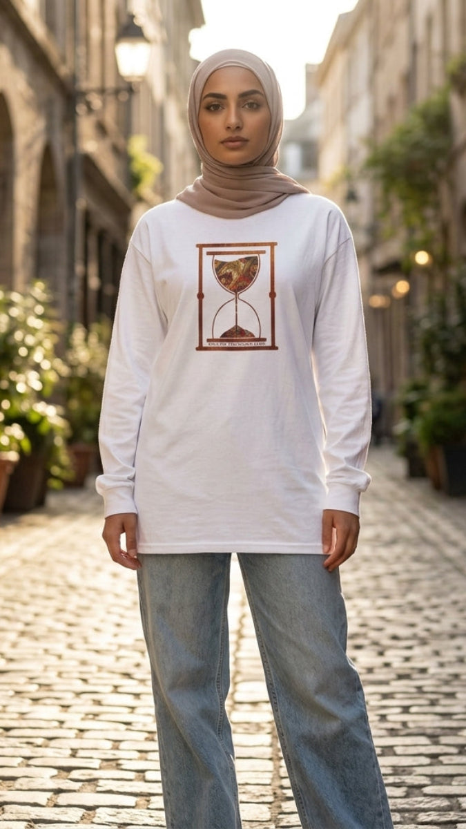 A woman wearing a white long-sleeve tunic with a brown hourglass graphic, paired with light blue jeans and a beige hijab, standing in a cobblestone alleyway.