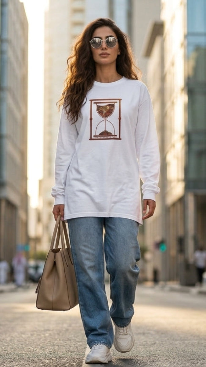 A woman walking down a city street wearing a white long-sleeve graphic t-shirt, light blue jeans, and white sneakers, carrying a tan tote bag.