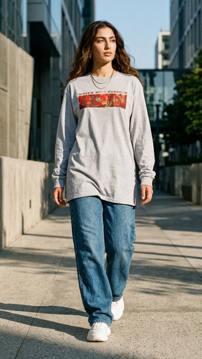 A woman walking down a sunlit urban sidewalk wearing a light gray oversized long-sleeve sweatshirt with a red graphic print across the chest, blue jeans, and white sneakers.