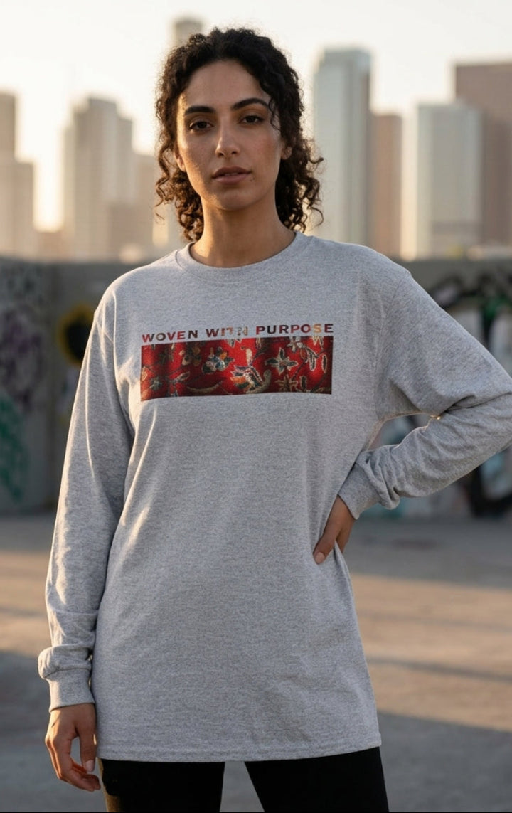 A woman wearing a light heather gray long-sleeve t-shirt with a red graphic print that reads 'WOVEN WITH PURPOSE' stands in an urban outdoor setting with graffiti walls and buildings in the background.