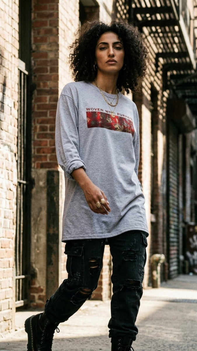 A woman stands on a city sidewalk wearing an oversized light gray long-sleeve graphic T-shirt with a red and gold rectangular chest print, paired with dark ripped jeans and black boots. The background shows brick buildings, fire escapes, and urban street details.