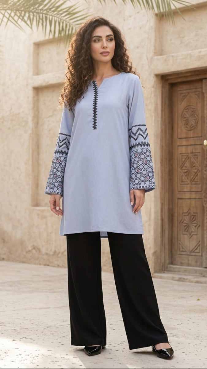 Woman in a light blue embroidered dress standing in front of a traditional building with a wooden door and palm tree.