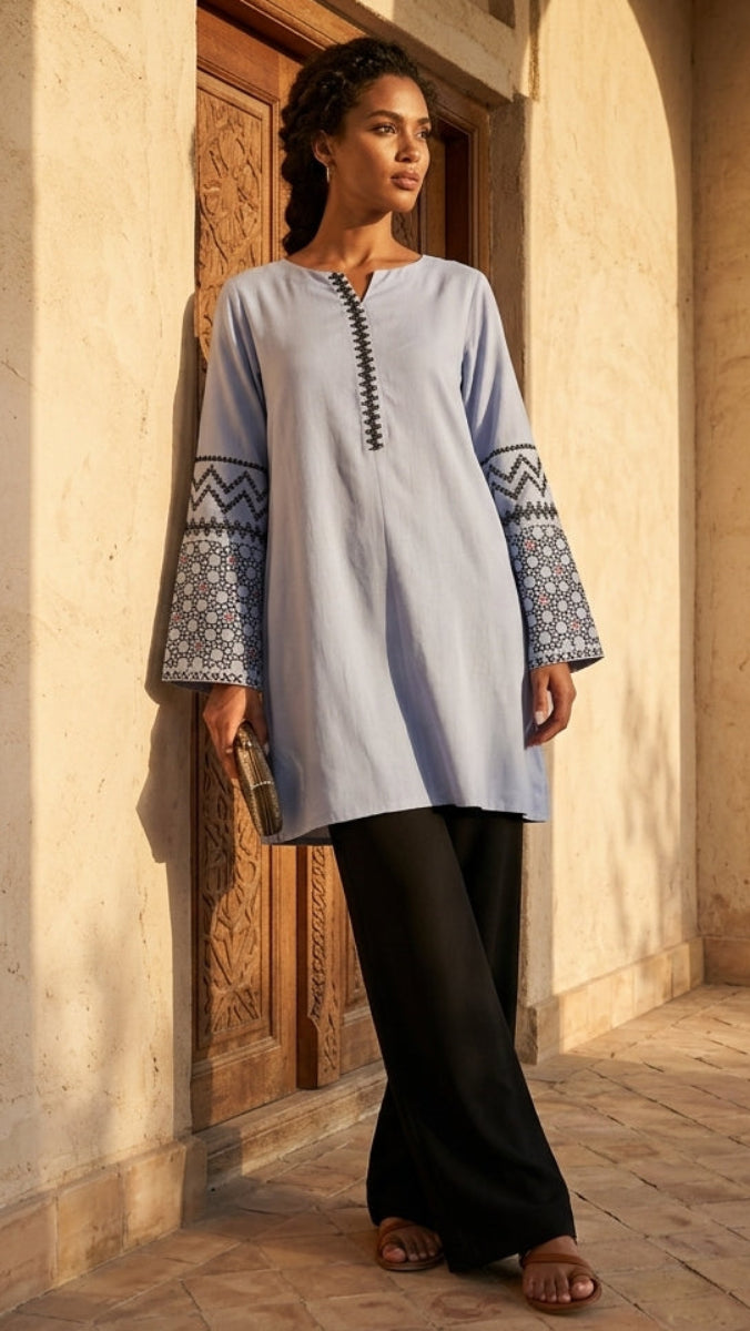 Woman wearing a light blue embroidered dress standing in a sunlit courtyard.