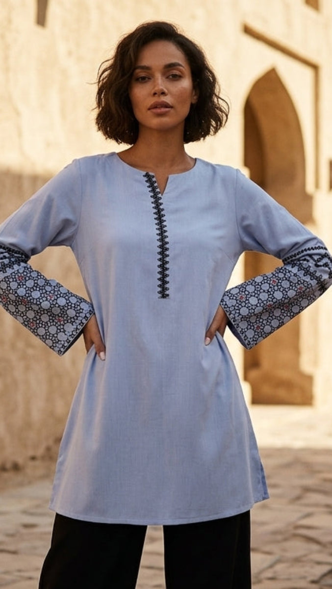 Woman wearing a light blue dress with patterned sleeves in an outdoor setting with stone architecture and palm trees.