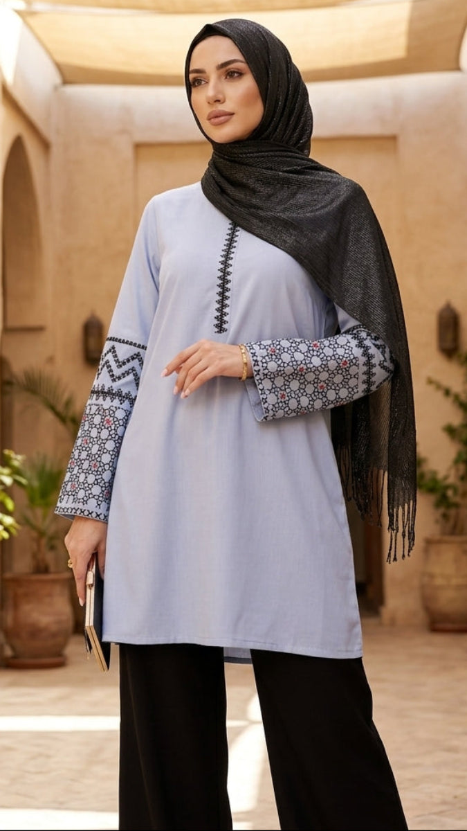 Woman in a light blue top and black hijab standing in a sunlit courtyard with arches and plants.