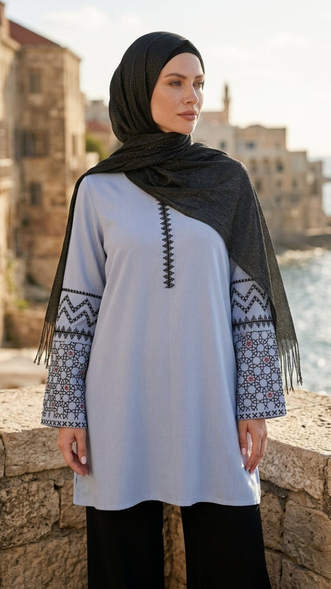 Woman wearing a white tunic with patterned sleeves and black hijab standing by a waterfront with buildings in the background.