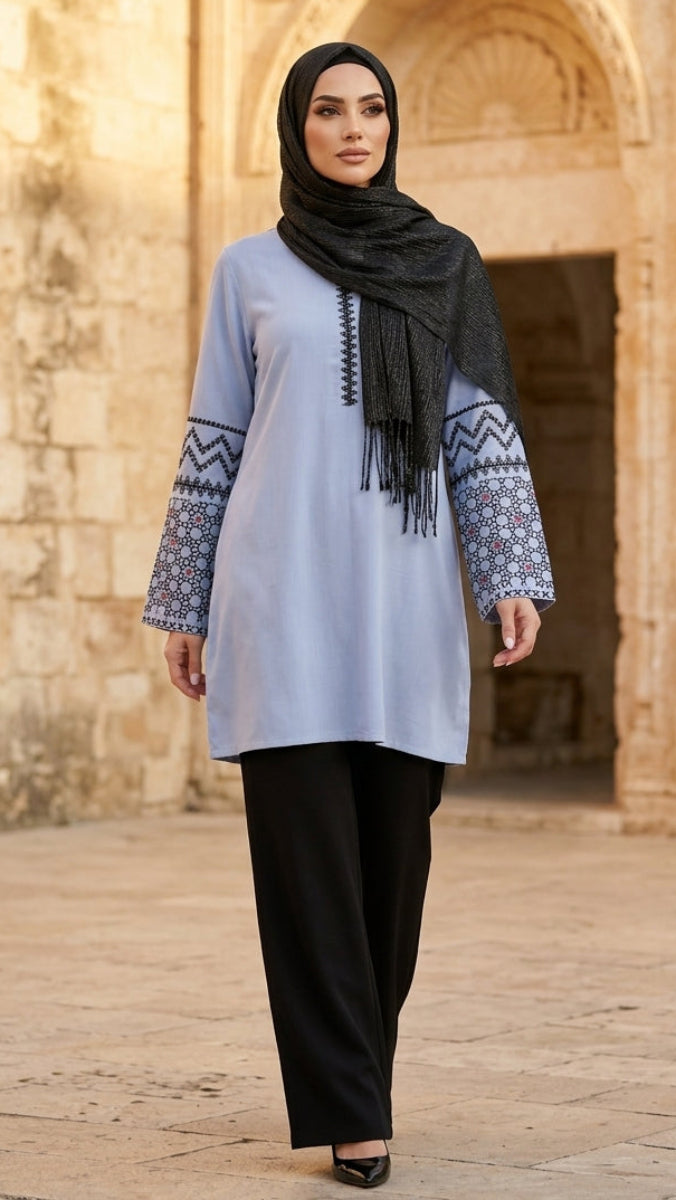 Woman wearing a light blue dress with patterned sleeves and black hijab in front of a stone building.
