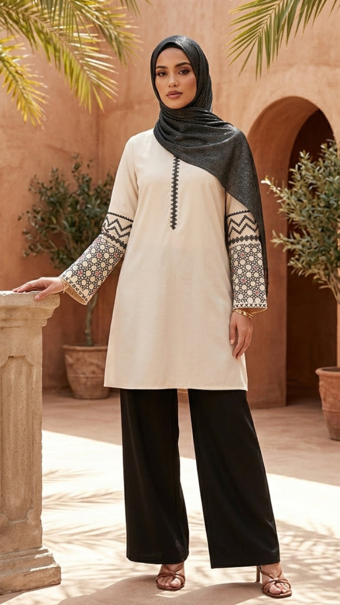 Woman in a cream and black outfit standing in a sunlit courtyard with arches and potted plants.