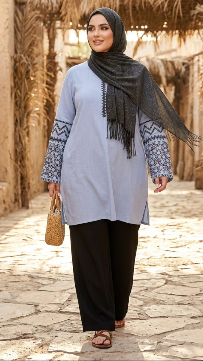 Woman in a light blue dress with patterned sleeves and black hijab standing in a sunlit alleyway.