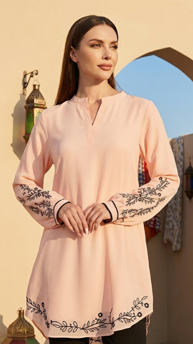 Woman wearing a light pink long-sleeve top with black embroidery in an outdoor setting.
