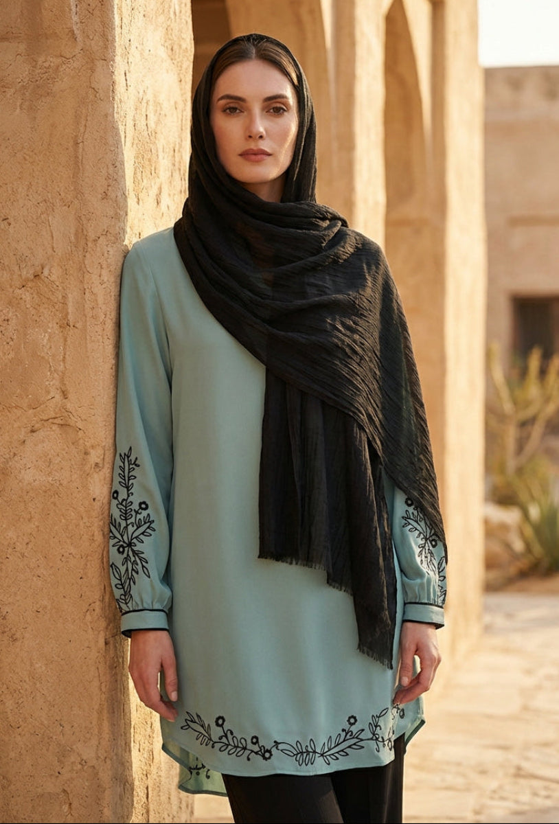 Woman wearing a light blue dress with black embroidery and a black hijab standing against a beige wall.