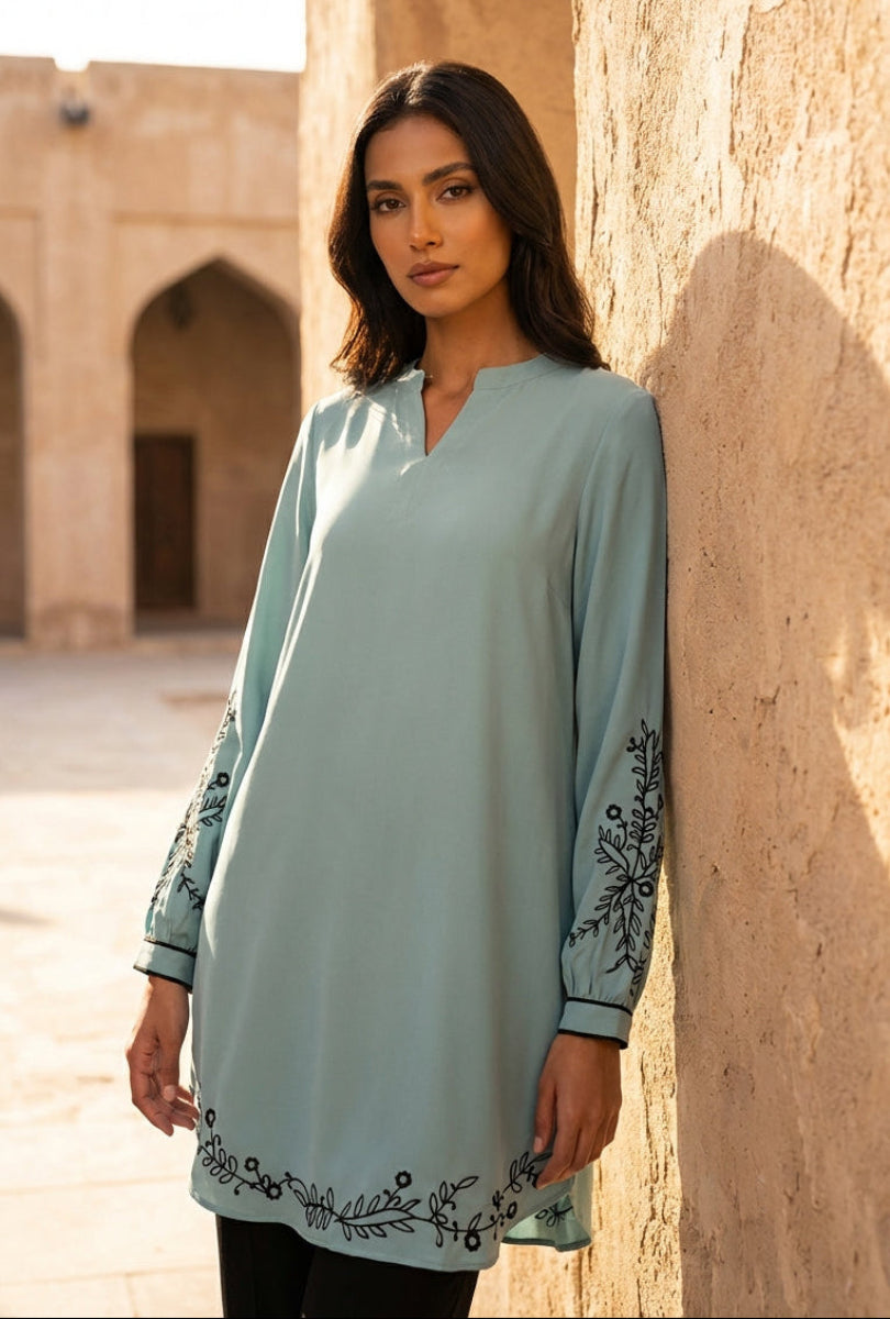 Woman wearing a light blue dress with embroidery, standing against a stone wall.