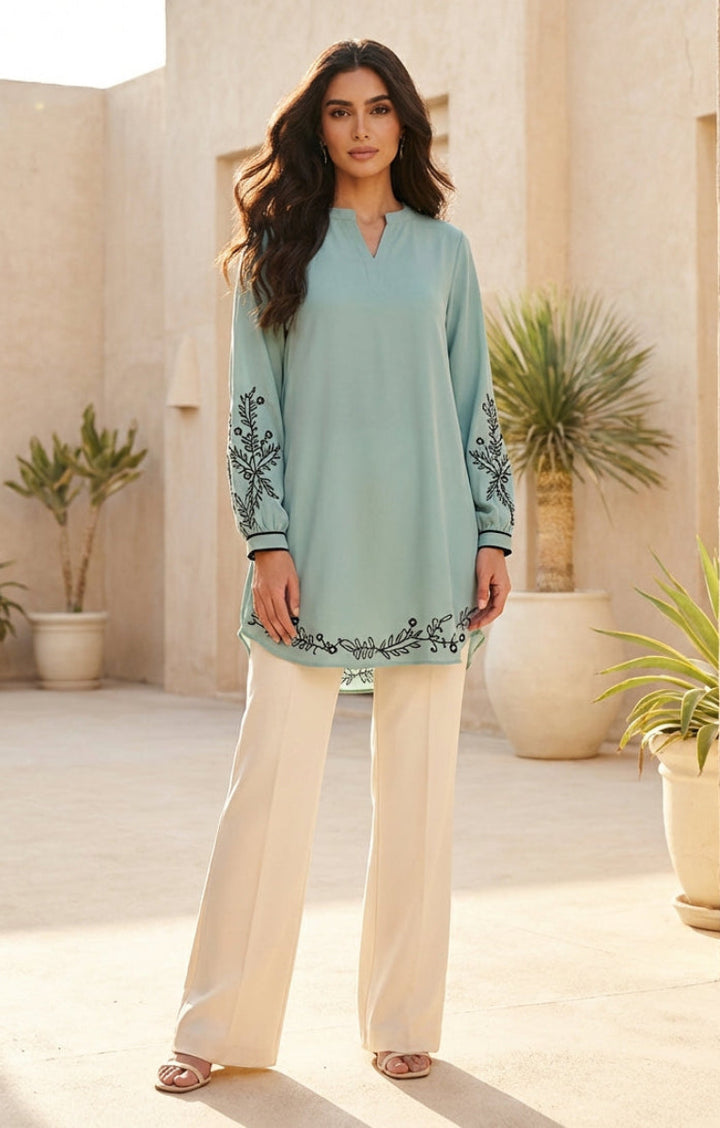 Woman wearing a light blue embroidered top and beige pants in an outdoor setting with plants and stone architecture.