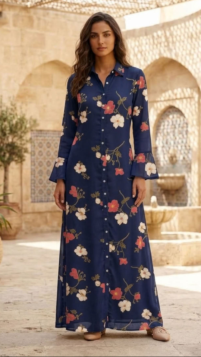 A woman stands in a sunlit courtyard wearing a long navy maxi dress with a white, pink, and red floral print. The setting features beige stone arches, potted plants, and a woven canopy overhead.