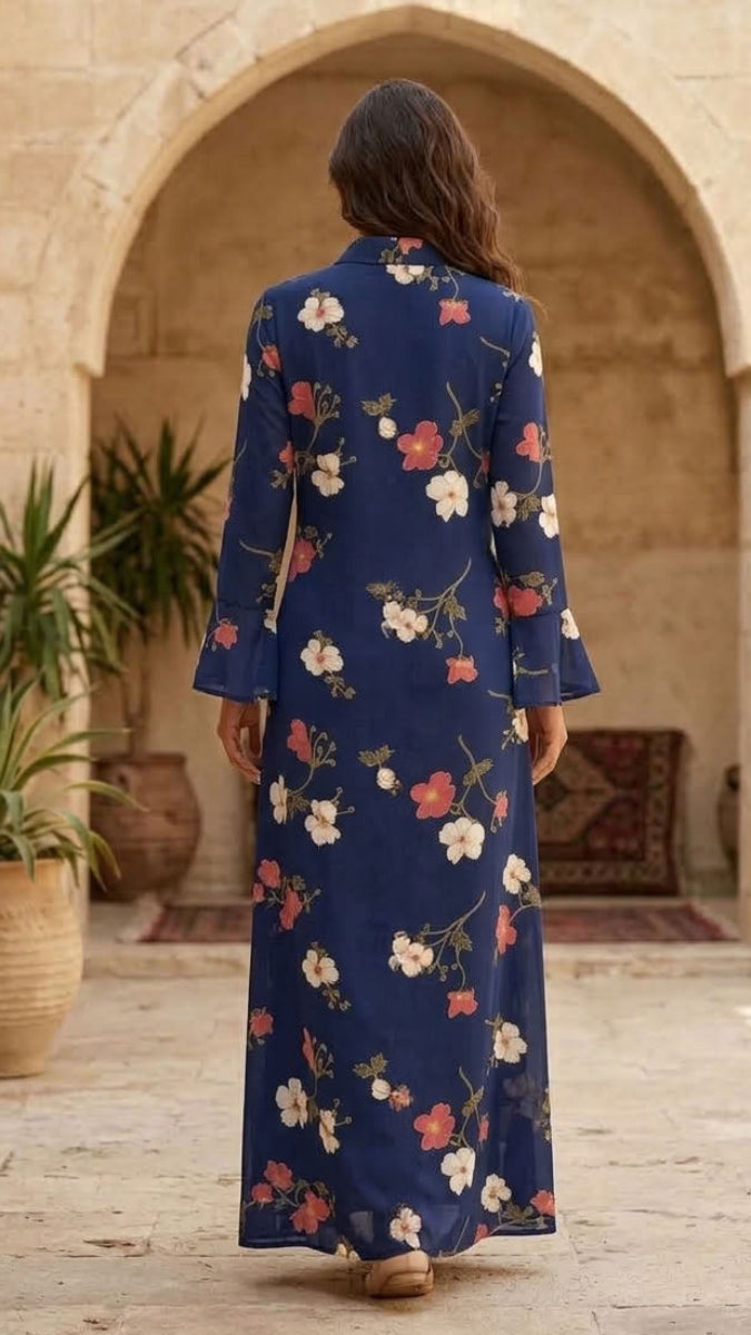 A woman viewed from behind wearing a navy blue long-sleeve maxi dress with a white, pink, and green floral pattern, standing in a stone courtyard with arches and potted plants.
