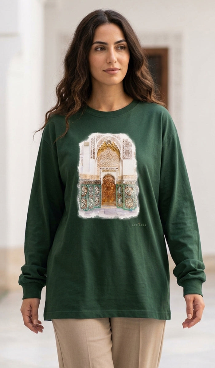A woman wearing a dark green long-sleeve t-shirt with a detailed architectural archway graphic on the front, paired with beige pants, standing in a brightly lit indoor setting.
