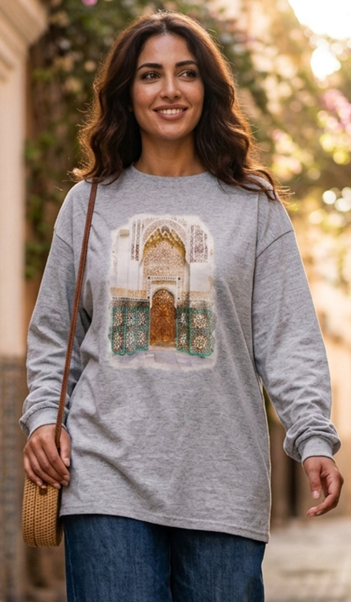 A woman wearing a heather gray long-sleeve t-shirt with a detailed graphic print of an Islamic archway (mihrab) on the front, paired with blue jeans and carrying a woven straw bag.
